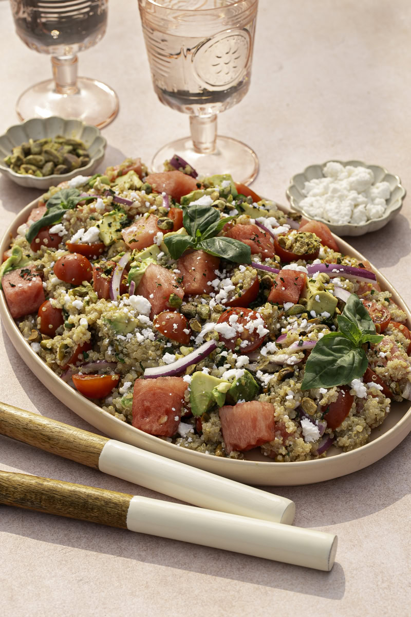 Watermelon Quinoa Salad on a serving plate with a bowl of feta cheese and a bowl of pistachios, two glasses of water, and salad servers.