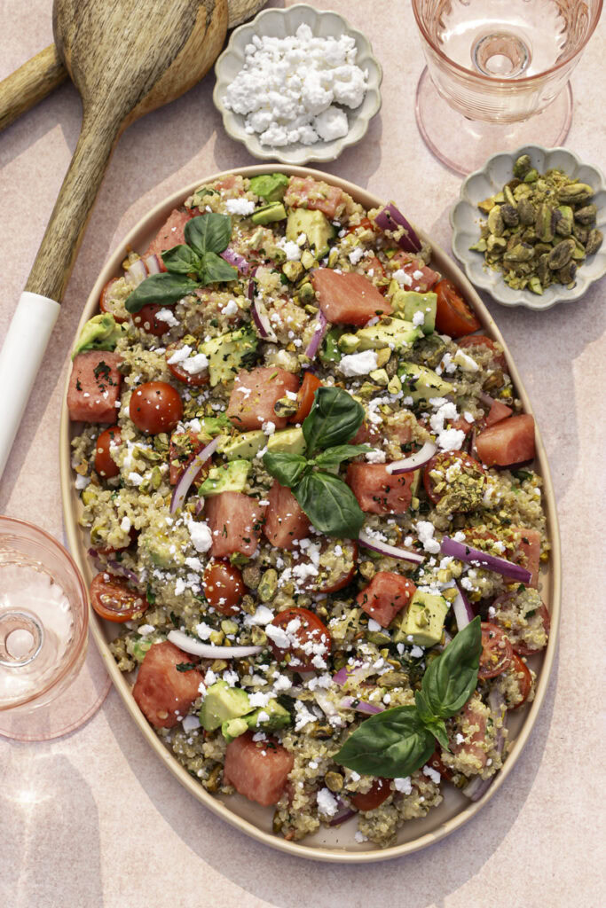 Watermelon Quinoa Salad on a serving plate with a bowl of feta cheese and a bowl of pistachios, two glasses of water, and salad servers.