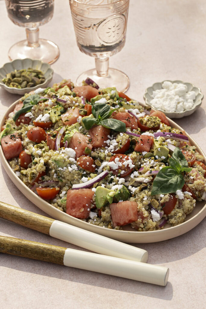 Watermelon Quinoa Salad on a serving plate with a bowl of feta cheese and a bowl of pistachios, two glasses of water, and salad servers.