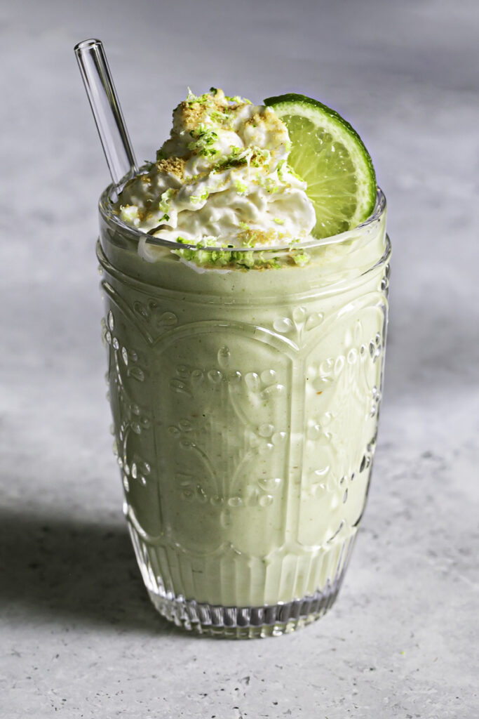a glass of key lime pie smoothie topped with dairy-free whipped cream garnished with graham cracker crumbs and lime zest.