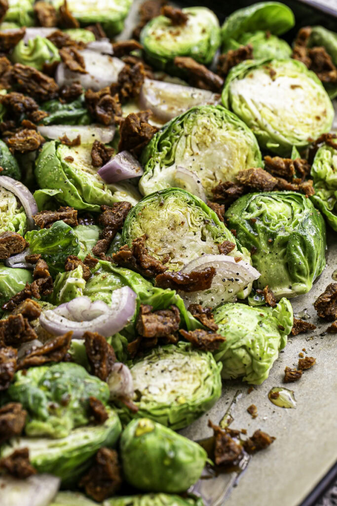 raw brussels sprouts combined with raw shallot and vegan bacon bits on a baking sheet before roasting.