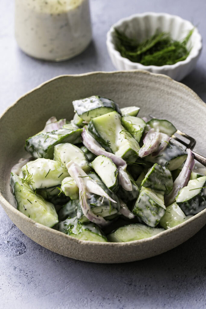 easy dairy-free creamy cucumber salad in a bowl with a jar of dressing and a bowl of fresh dill in the background.