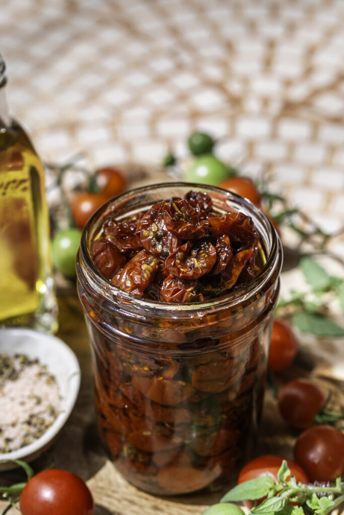 a jar of sun-dried cherry tomatoes in olive oil surrounded by fresh cherry tomatoes, a little jar of olive oil, and a small bowl of salt and herbs.