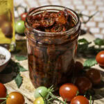 a jar of sun-dried cherry tomatoes in olive oil surrounded by fresh cherry tomatoes, a little jar of olive oil, and a small bowl of salt and herbs.