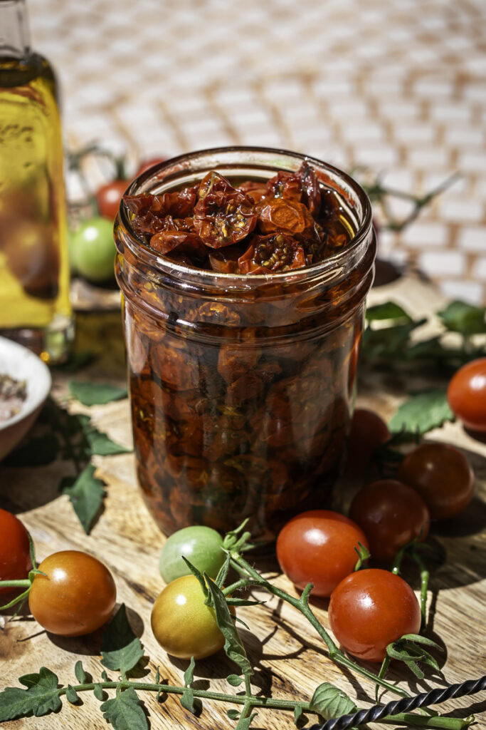 a jar of sun-dried cherry tomatoes in olive oil surrounded by fresh cherry tomatoes, a little jar of olive oil, and a small bowl of salt and herbs.