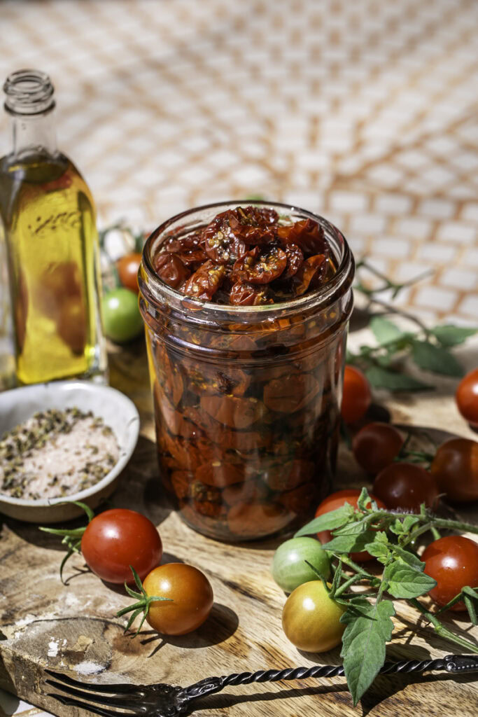 a jar of sun-dried cherry tomatoes in olive oil surrounded by fresh cherry tomatoes, a little jar of olive oil, and a small bowl of salt and herbs.