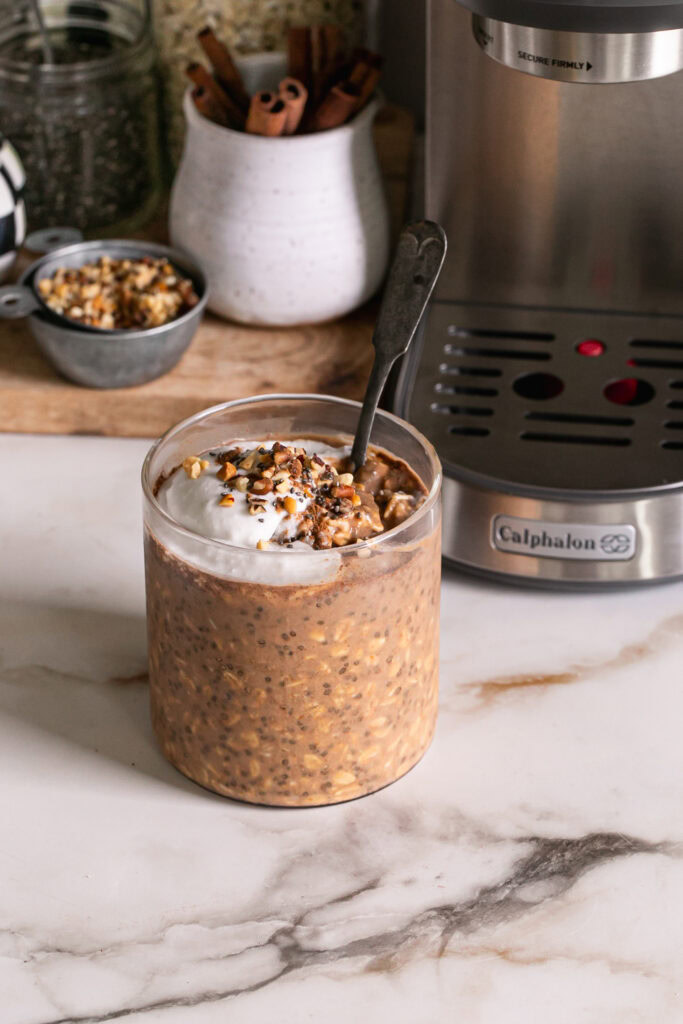 pumpkin spice espresso overnight oats in a glass with a spoon topped with coconut cream, and chopped nuts.