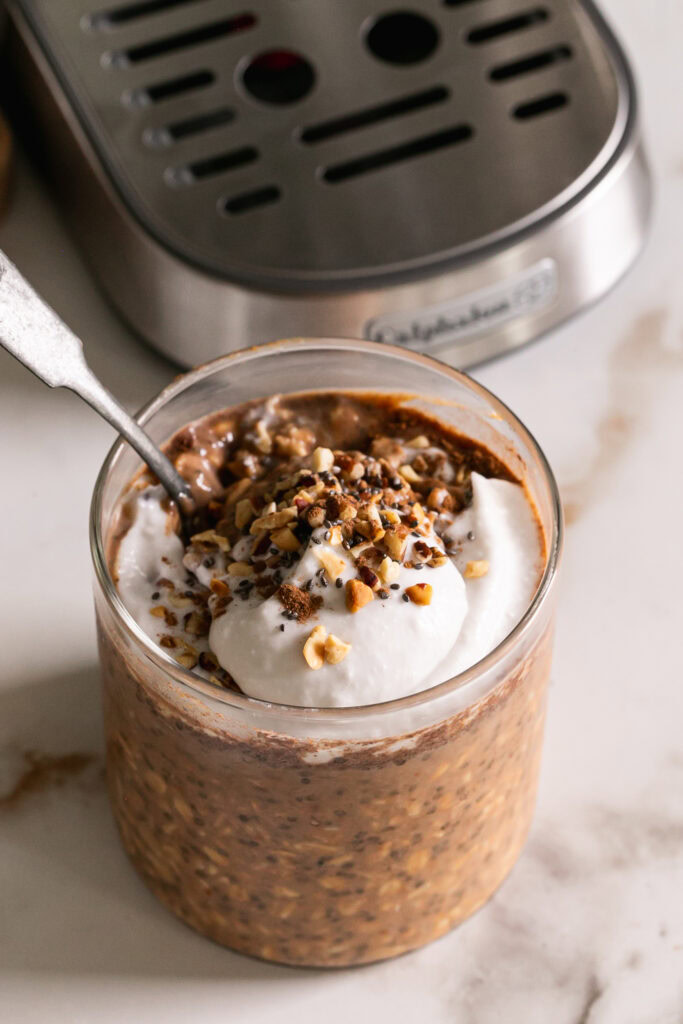 overhead shot of pumpkin spice espresso overnight oats in a glass with a spoon topped with coconut cream, and chopped nuts.