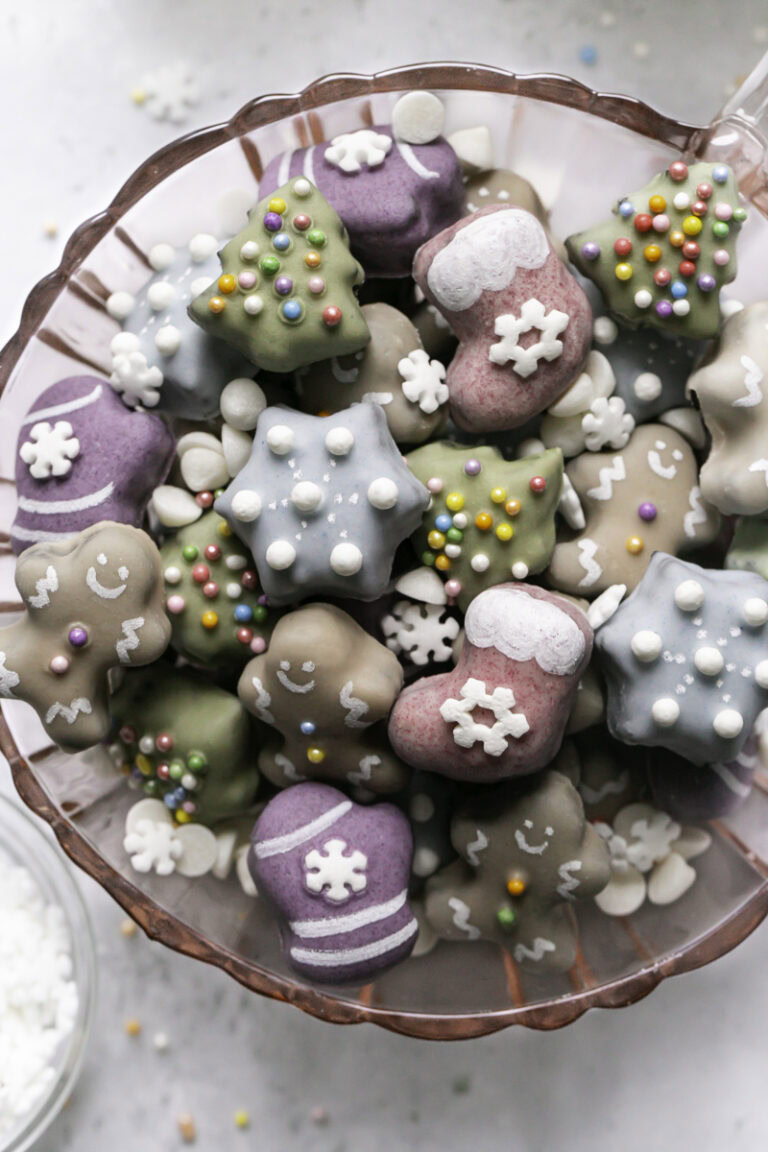 Close up of Mini Chocolate Almond Flour Christmas Cookies in a pink glass mug with sprinkles, white chocolate chips and candy snowflakes sprinkled all around.