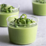 three glasses creamy green gazpacho garnished with cucumber ribbons, basil leaves and peas.