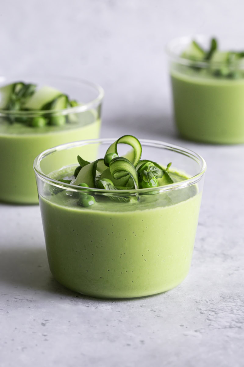 three glasses creamy green gazpacho garnished with cucumber ribbons, basil leaves and peas.