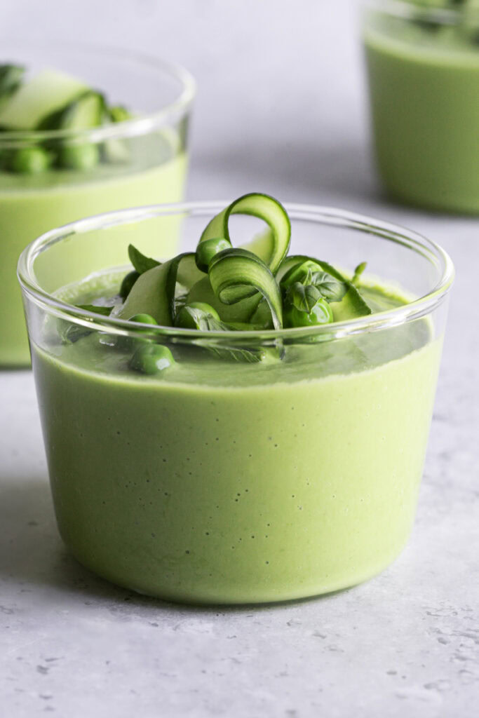 a close up of a glass of creamy green gazpacho garnished with cucumber ribbons, basil leaves and peas.