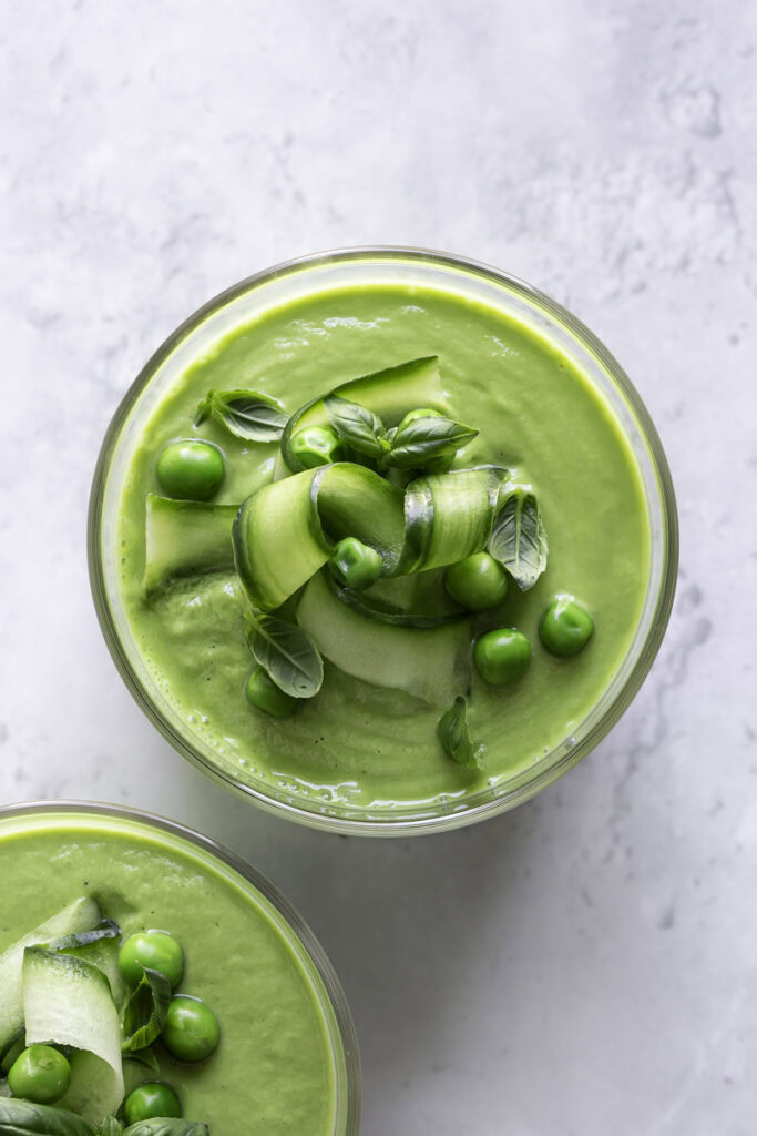 overhead shot of a glass of creamy green gazpacho garnished with cucumber ribbons, basil leaves and peas.