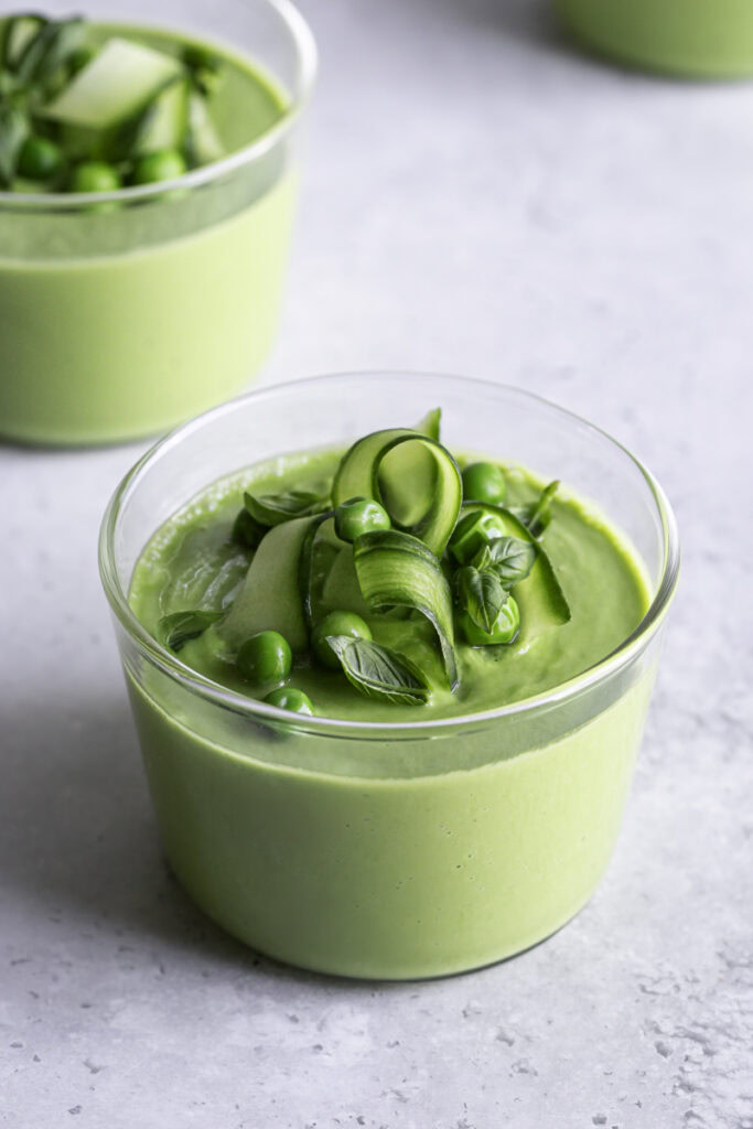 3/4 shot of creamy green gazpacho garnished with cucumber ribbons, basil leaves and peas.