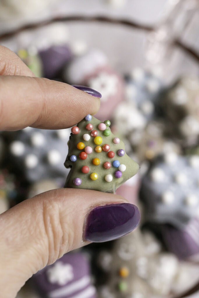 fingers holding a mini Christmas tree cookie with the rest of the cookies blurred in the background.