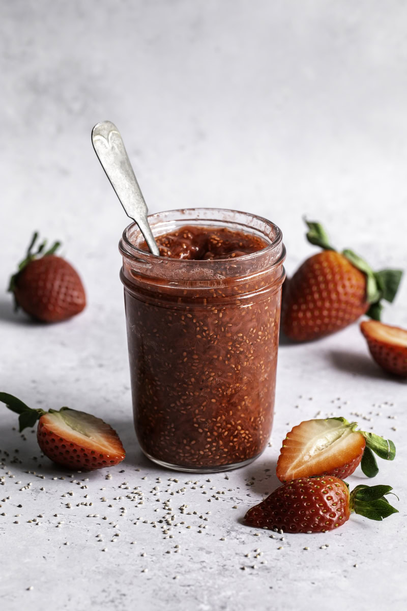 a jar of strawberry chia seed jam with a spoon in it and fresh strawberries around it.