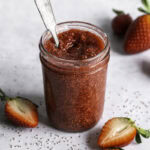 a jar of strawberry chia seed jam with a spoon in it and fresh strawberries around it.