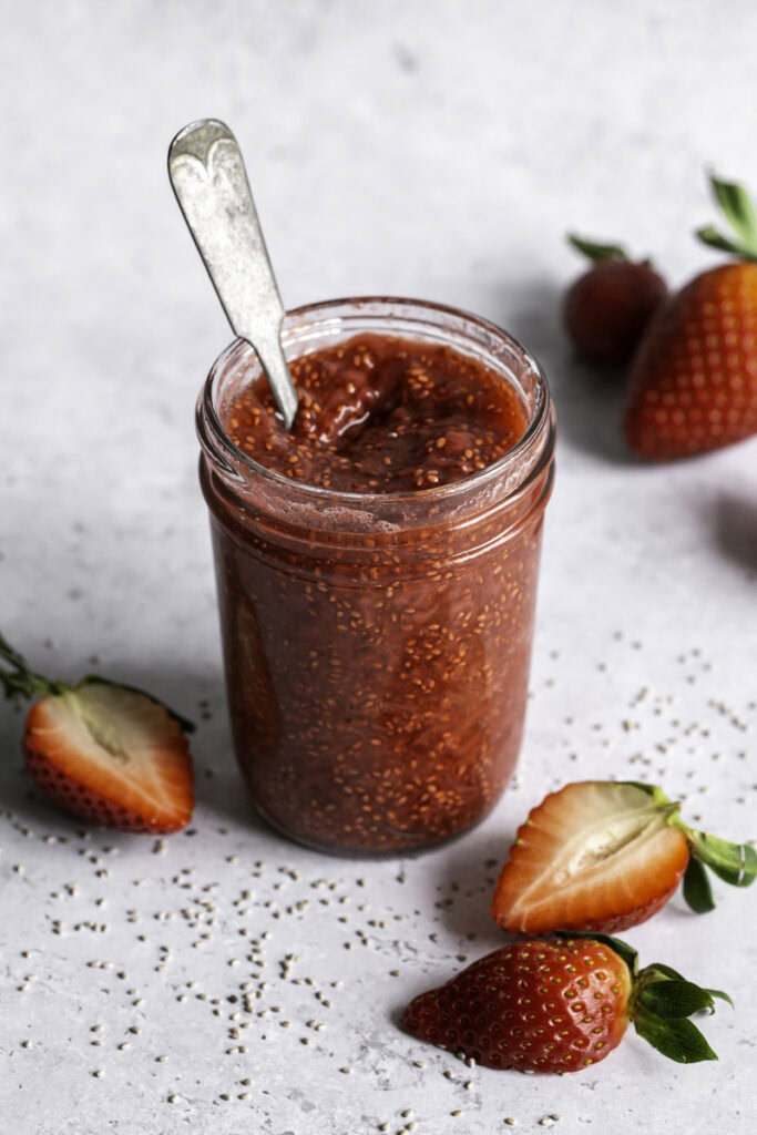 a jar of strawberry chia seed jam with a spoon in it and fresh strawberries around it.