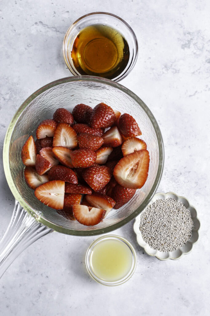 sliced strawberries in a glass saucepan with a bowl of maple syrup, a bowl of chia seeds, and a bowl of lemon juice beside it.