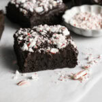 Fudgy Flourless Peppermint Date Brownies cut into squares on a piece of parchment paper with a bowl of crushed candy canes.