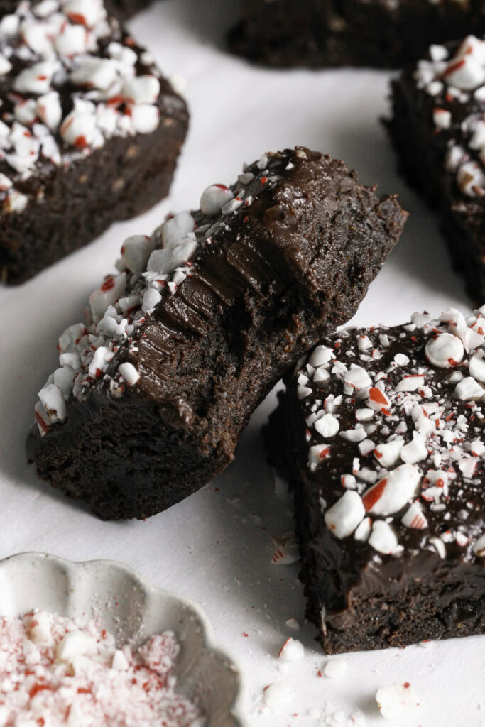 close up of a Fudgy Flourless Peppermint Date Brownies cut into squares, one with a bite taken out of it and a bowl of crushed candy canes.