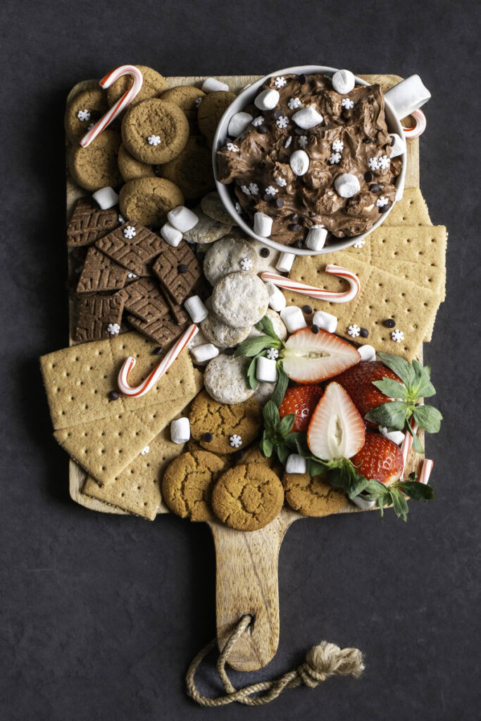 a board of cookies, graham crackers, strawberries, candy canes, and a mug of creamy vegan hot cocoa dessert dip.