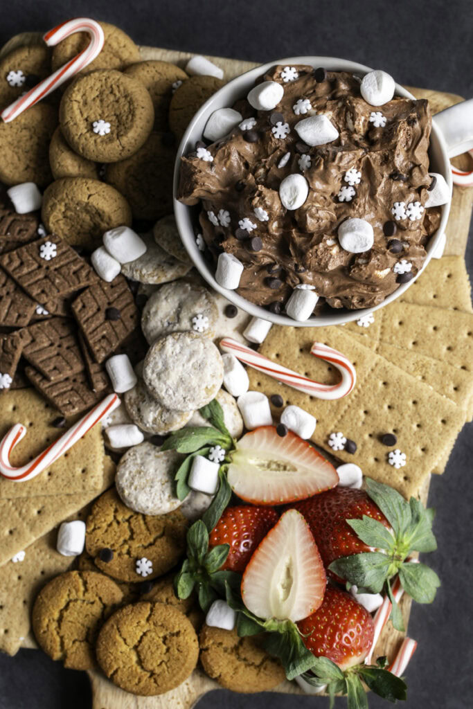 a board of cookies, graham crackers, strawberries, candy canes, and a mug of creamy vegan hot cocoa dessert dip.