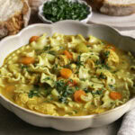 3/4 image of a big bowl of Vegan Lemon Turmeric Chicken Noodle Soup with slices of baked bread and a bowl of fresh chopped parsley.