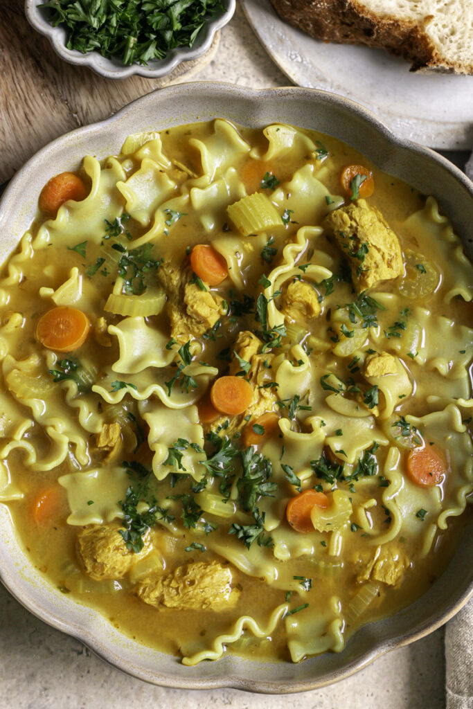 a close up of a big bowl of vegan lemon turmeric chicken noodle soup with a bowl of fresh parsley and a slice of baked bread.