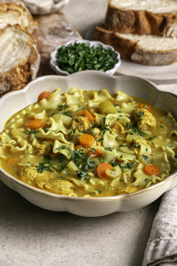 3/4 image of a big bowl of Vegan Lemon Turmeric Chicken Noodle Soup with slices of baked bread and a bowl of fresh chopped parsley.