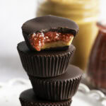 Peanut Butter & Jelly Chocolate Cups stacked on top of one another, the top one with a bite taken out of it to reveal the peanut butter and jelly filling.