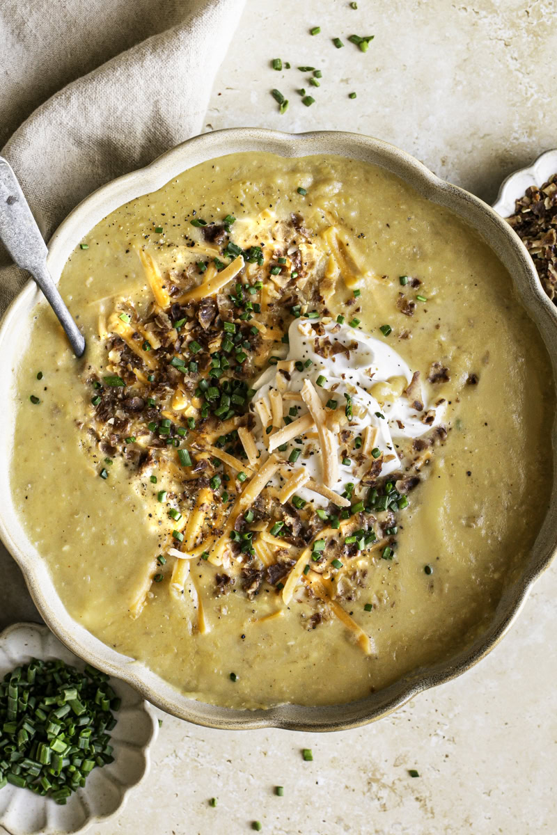 close up of a bowl of loaded baked potato soup with small bowls of chopped chives and vegan bacon bits.