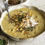 3/4 shot of a bowl of loaded baked potato soup with small bowls of chopped chives and vegan bacon bits.