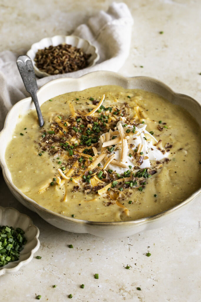 3/4 shot of a bowl of loaded baked potato soup with small bowls of chopped chives and vegan bacon bits.