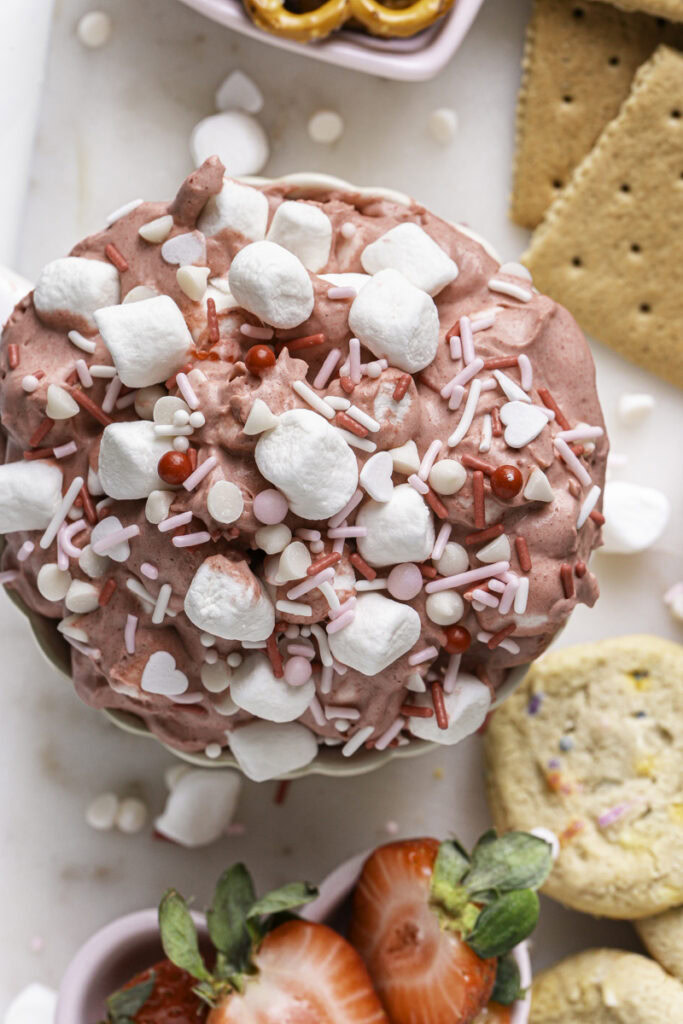 close up overhead of Strawberry White Chocolate Coconut Milk Marshmallow Dip with fresh strawberries, cookies, graham crackers, and pretzels around it.