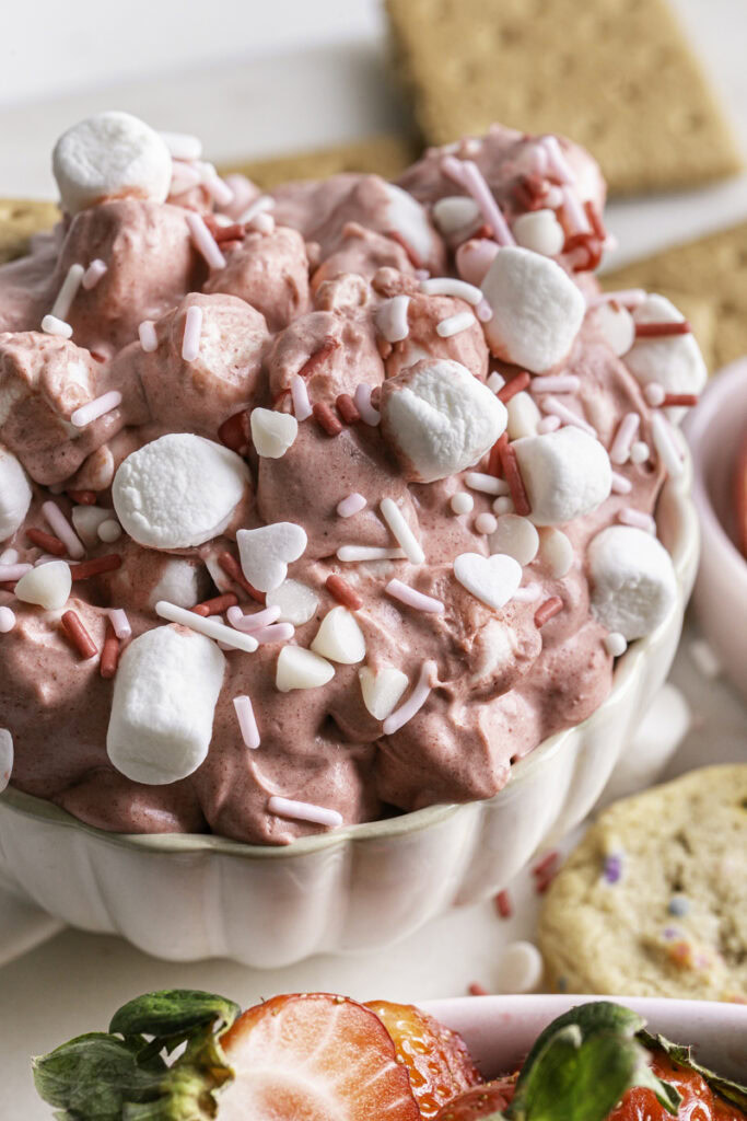 image of Strawberry White Chocolate Coconut Milk Marshmallow Dip in a mug surrounded by fresh strawberries, cookies, graham crackers, and pretzels.