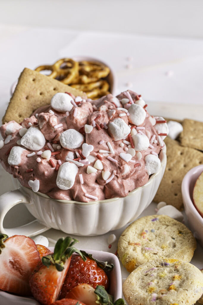 3/4 image of Strawberry White Chocolate Coconut Milk Marshmallow Dip in a mug surrounded by fresh strawberries, cookies, graham crackers, and pretzels.