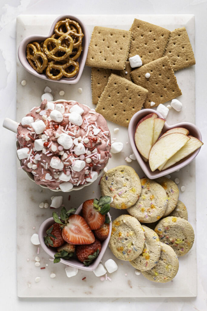 overhead shot of a dessert snack board with Strawberry White Chocolate Coconut Milk Marshmallow Dip, sprinkle cookies, apple slices, strawberries, graham crackers, and pretzels.