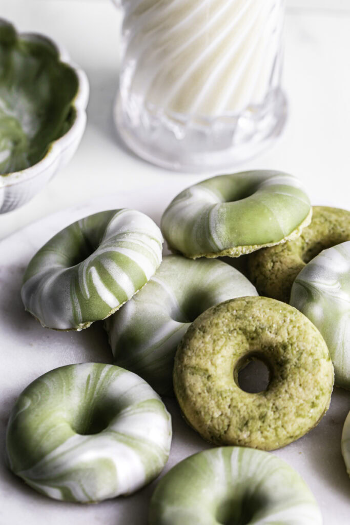 No-Bake Protein Matcha Donuts piled on top of one another on a round marble plate with melted matcha white chocolate and milk in the background.