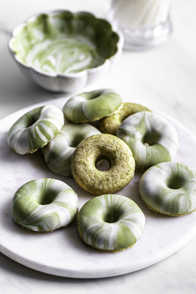 No-Bake Protein Matcha Donuts piled on top of one another on a round marble plate with melted matcha white chocolate and milk in the background.