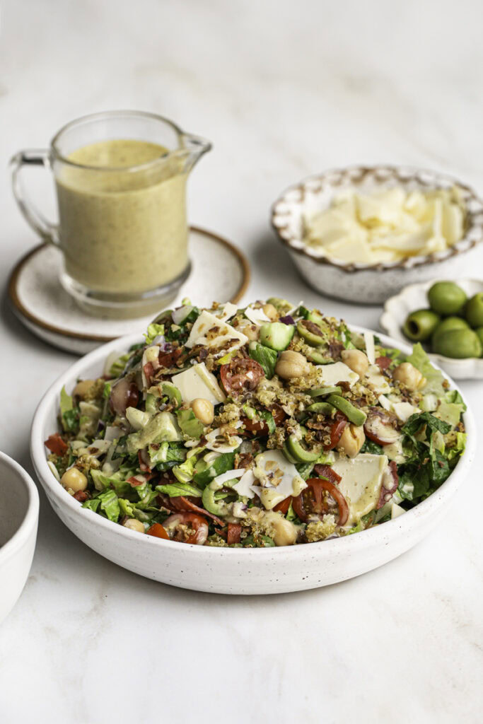 Vegan Italian Chopped Salad with Herby Crispy Quinoa in a bowl with a jar of salad dressing, a bowl of vegan parmesan cheese, and a bowl of green olives in the background.