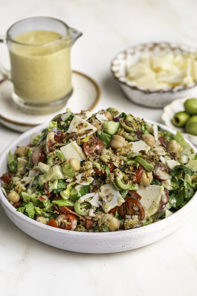 Vegan Italian Chopped Salad with Herby Crispy Quinoa in a bowl with a jar of salad dressing, a bowl of vegan parmesan cheese, and a bowl of green olives in the background.