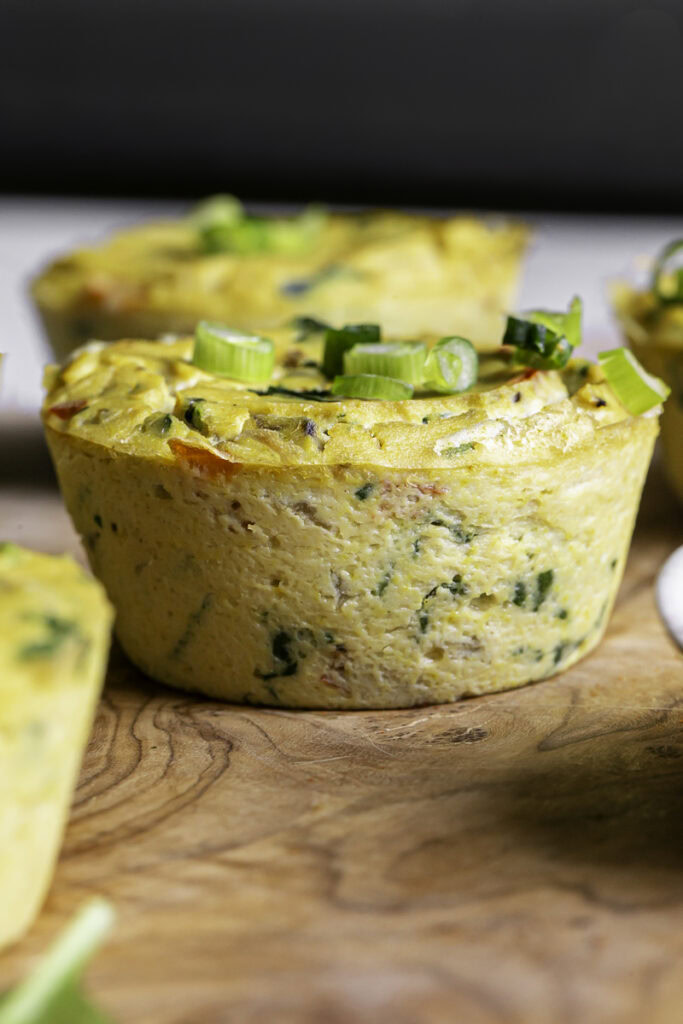 a close up of a vegan baked spinach mushroom pepper onion egg bite on a wooden cutting board garnished with sliced green onions.