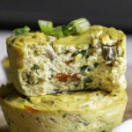 two vegan baked spinach mushroom pepper onion egg bites on a wooden cutting board, the top one with a bite taken out of it to reveal the soft egg-like texture.