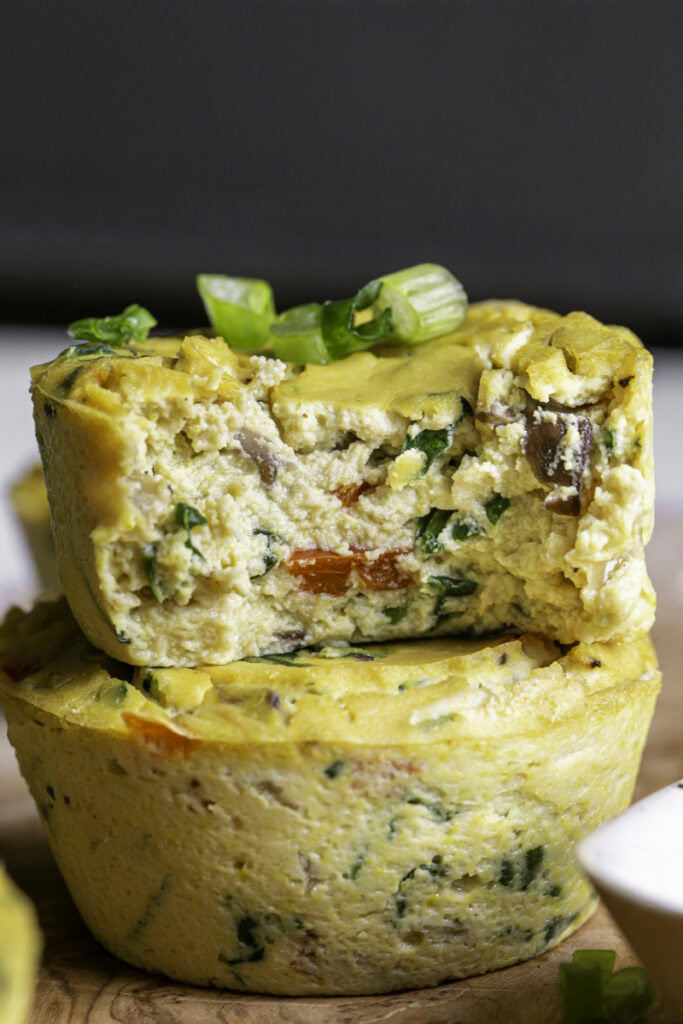 two vegan baked spinach mushroom pepper onion egg bites on a wooden cutting board, the top one with a bite taken out of it to reveal the soft egg-like texture.