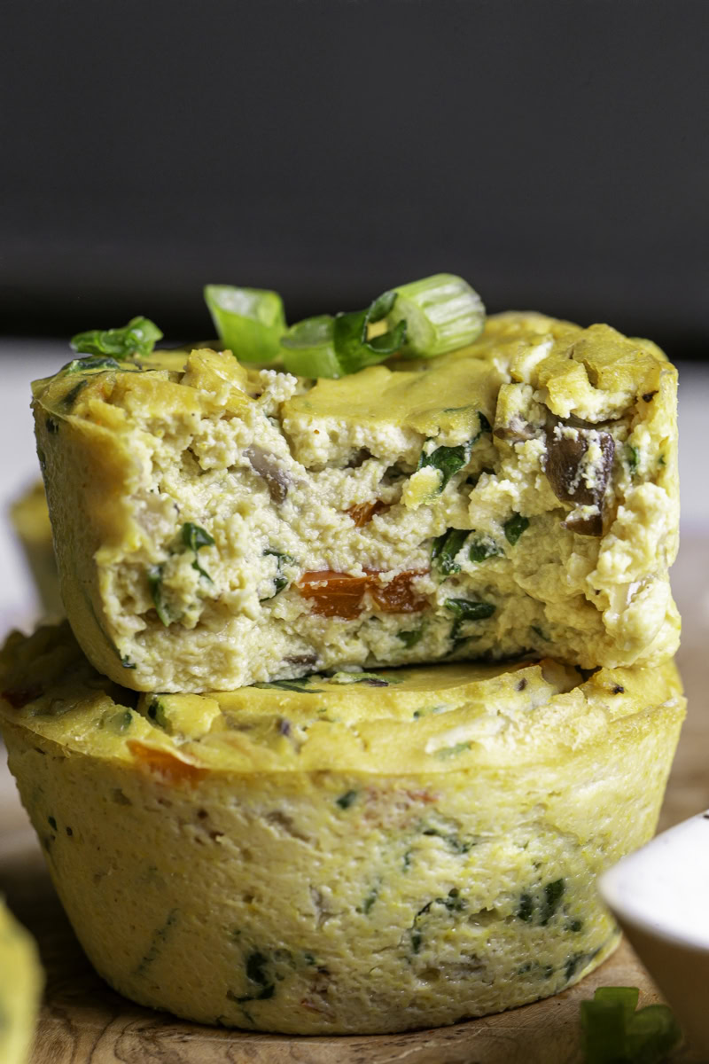two vegan baked spinach mushroom pepper onion egg bites on a wooden cutting board, the top one with a bite taken out of it to reveal the soft egg-like texture.