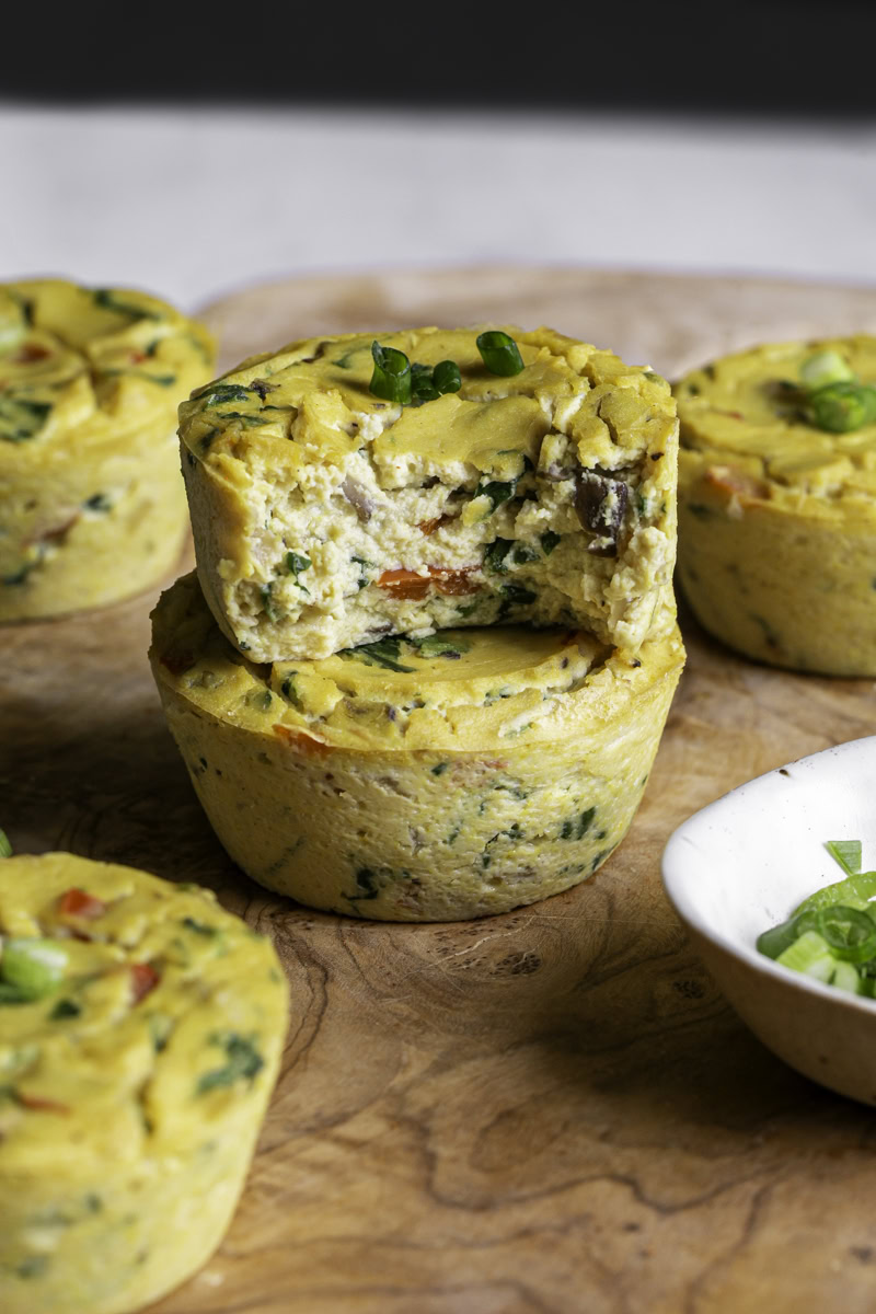 two vegan baked spinach mushroom pepper onion egg bites on a wooden cutting board, the top one with a bite taken out of it to reveal the soft egg-like texture.