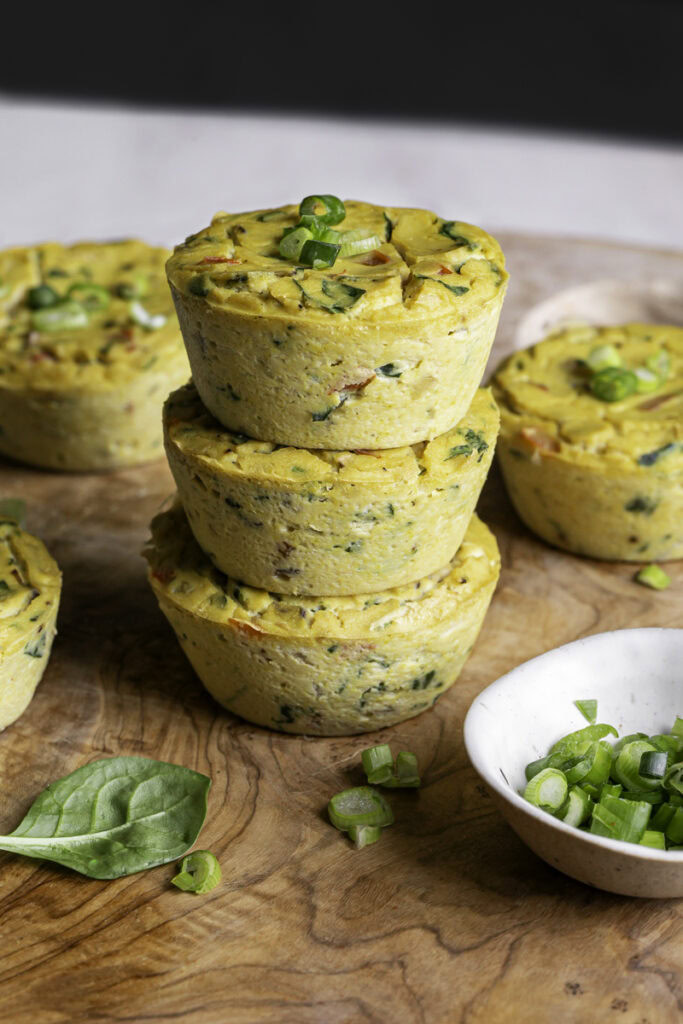 three vegan baked spinach mushroom pepper onion egg bites stacked on top of one another on a wooden cutting board.