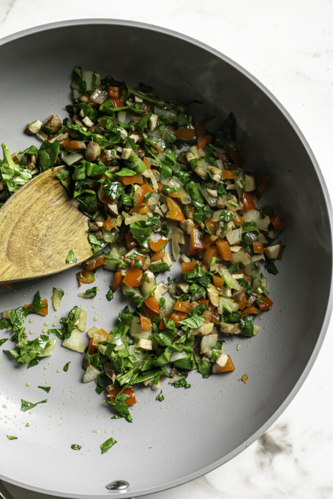 chopped spinach tossed into the skillet with the onion, mushrooms, and red bell pepper with a wooden spoon in the skillet.
