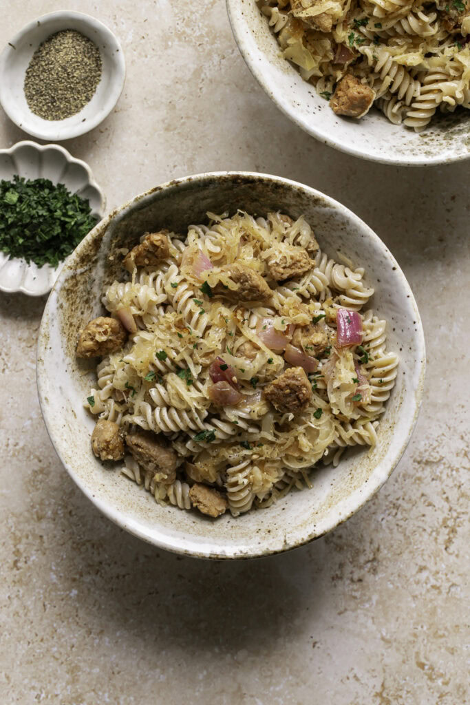 two bowls of vegan Bratwurst & Sauerkraut Pasta, a small bowl of chopped parsley and black pepper.